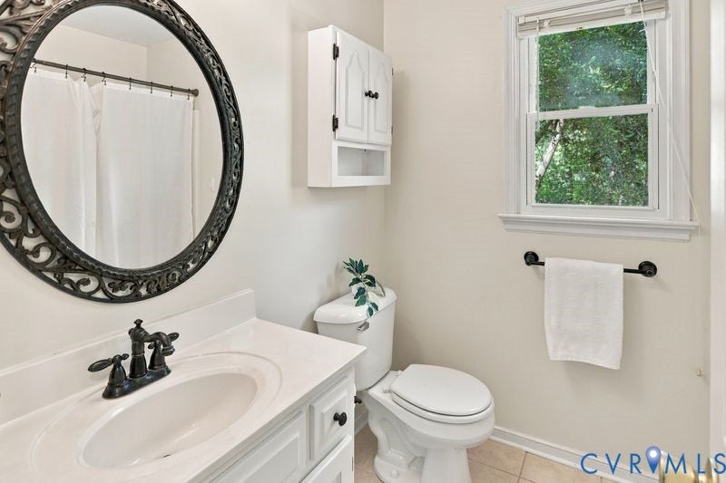 10208 Claybar Trail, Chesterfield, VA 23236 White vanity featuring a smooth-surface countertop, integrated oval basin, and matte black widespread faucet, paired with a decorative circular framed mirror, wall-mounted cabinet, and window with natural light