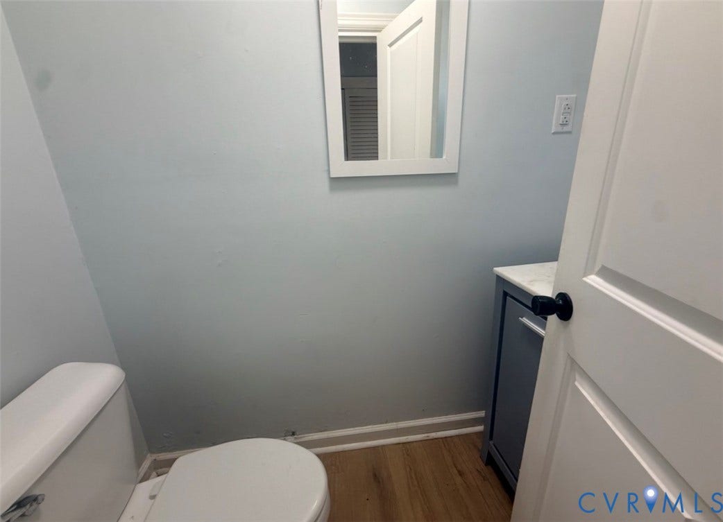 Bathroom featuring wood-finish flooring, a charcoal-toned vanity cabinet with a stone-top finish, a wall-mounted rectangular mirror, and a white toilet