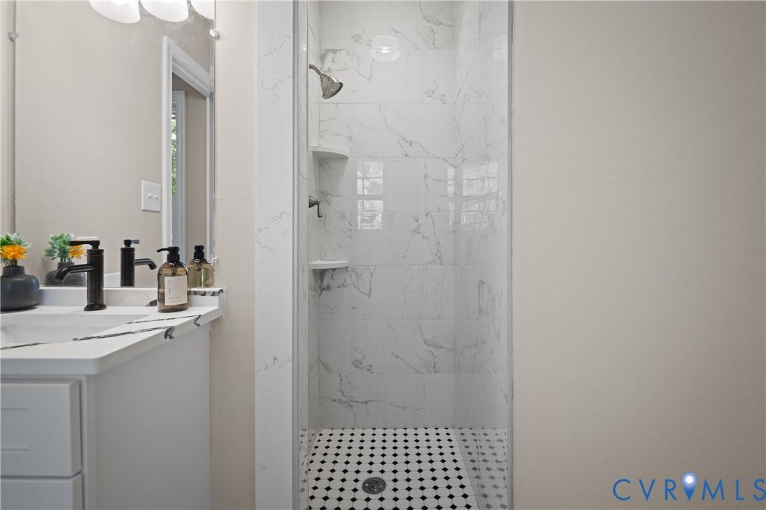 9010 Fawn Lane, Henrico, VA 23229 Shower stall featuring large-format veined tile surround and built-in shelving, complemented by a mosaic tile floor, white vanity with stone-finish countertop, and dark-finish faucet fixtures