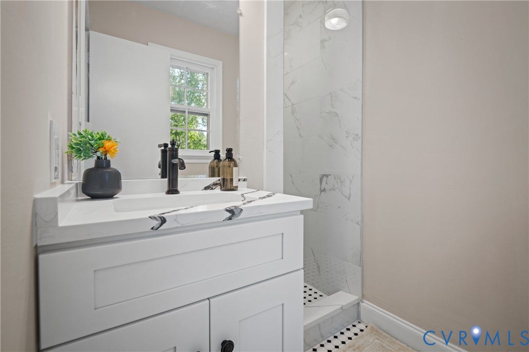 9010 Fawn Lane, Henrico, VA 23229 White vanity with an integrated basin and stone-patterned countertop featuring a matte black faucet, paired with a shower enclosure showcasing large-format surround tile and a geometric mosaic floor