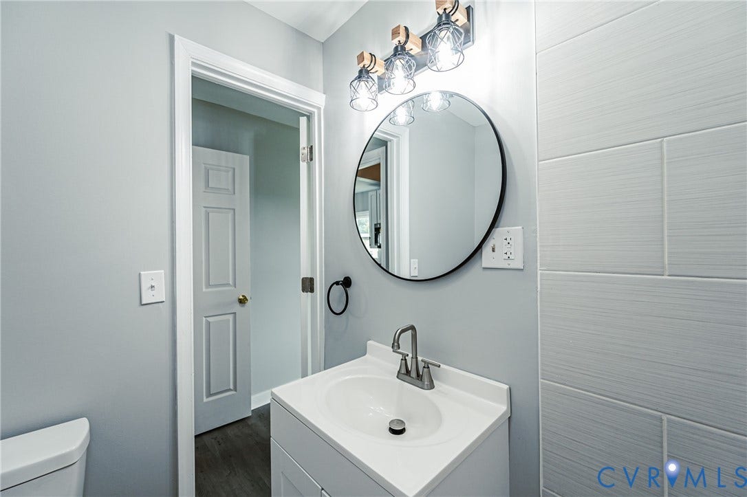Bathroom vanity featuring a solid-surface integrated sink, circular black-framed mirror, three-light industrial-style fixture, and horizontal textured wall tiling