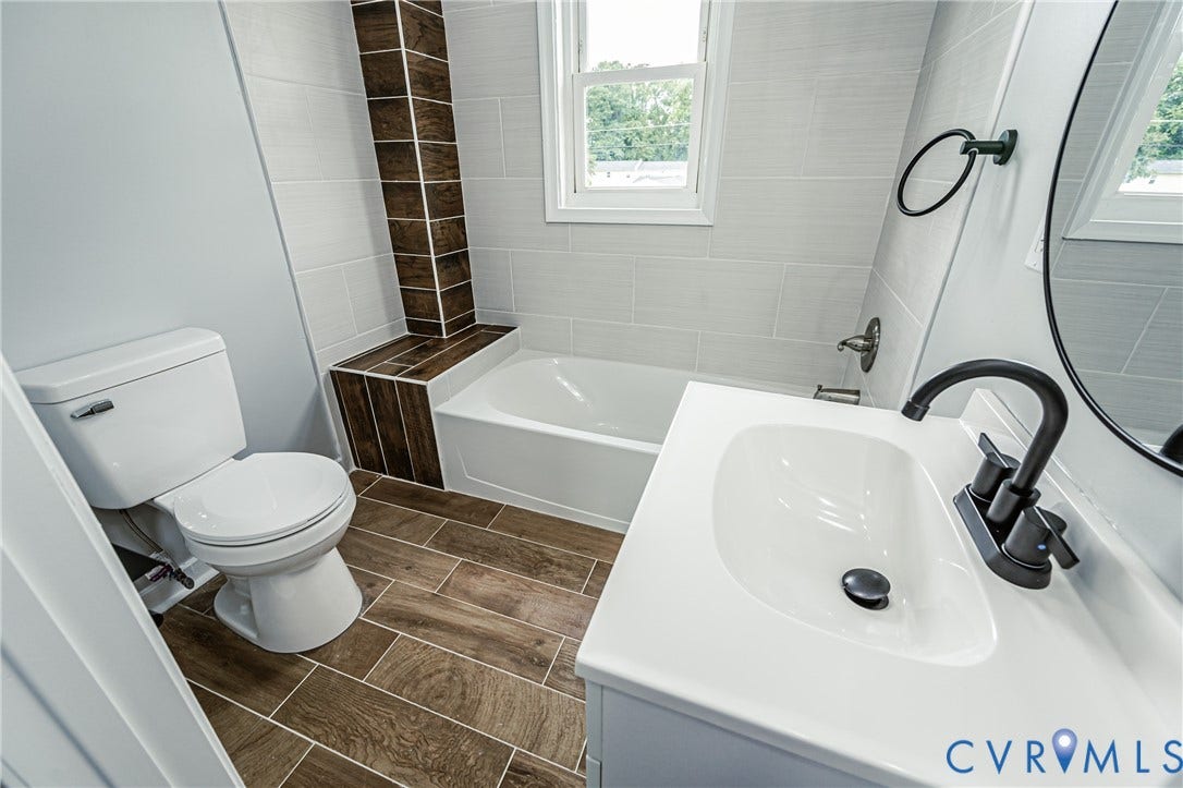 Bathroom featuring wood-finish floor tiling, a white vanity with a composite countertop, matte black hardware, a tub with integrated tile surround, and wood-finish decorative wall accents