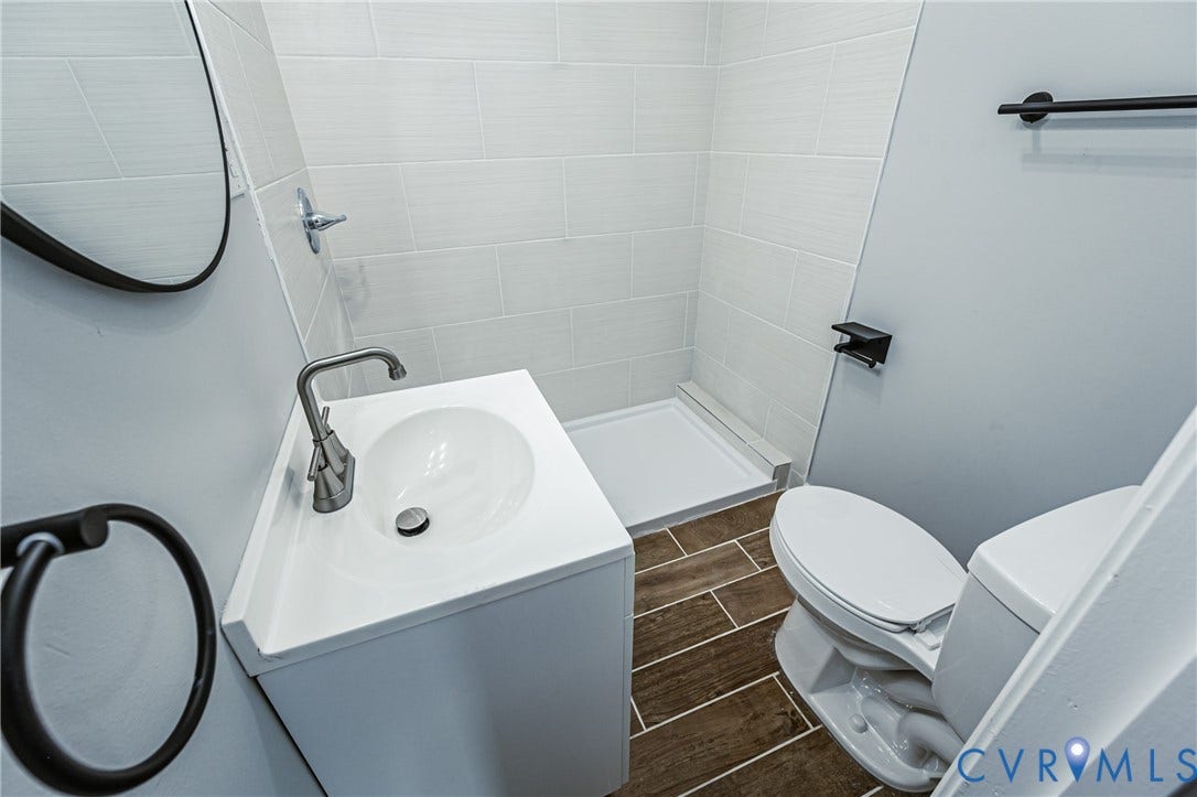 White vanity featuring an integrated sink with a brushed-finish faucet, walk-in shower with horizontal tile surround, wood-finish flooring, and matte black hardware