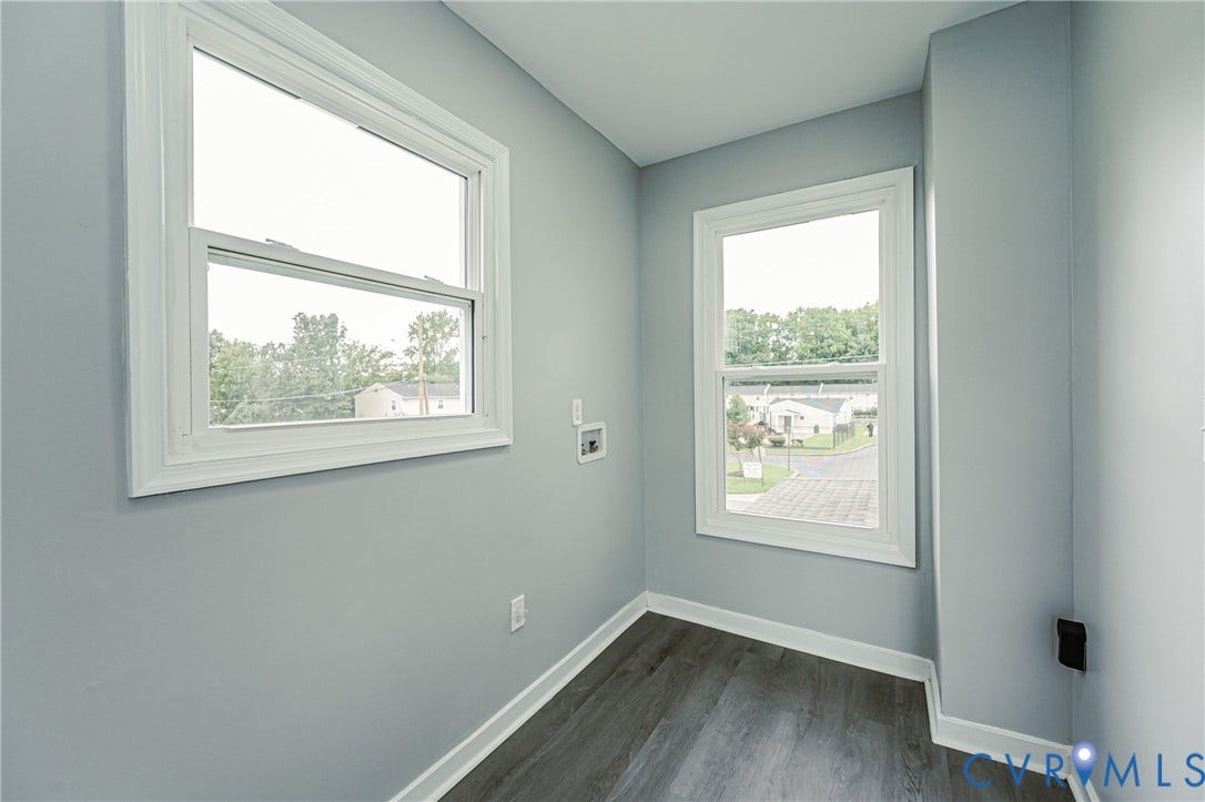 Neutral-toned interior space featuring wood-finish flooring, two double-hung windows, and utility hookups