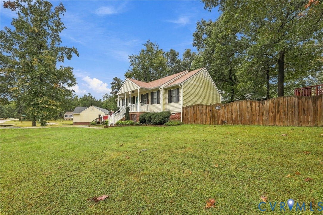 Exterior featuring light-toned horizontal siding, a covered front porch with white railing, and a brick foundation, situated on a lot with expansive grass coverage and mature perimeter trees