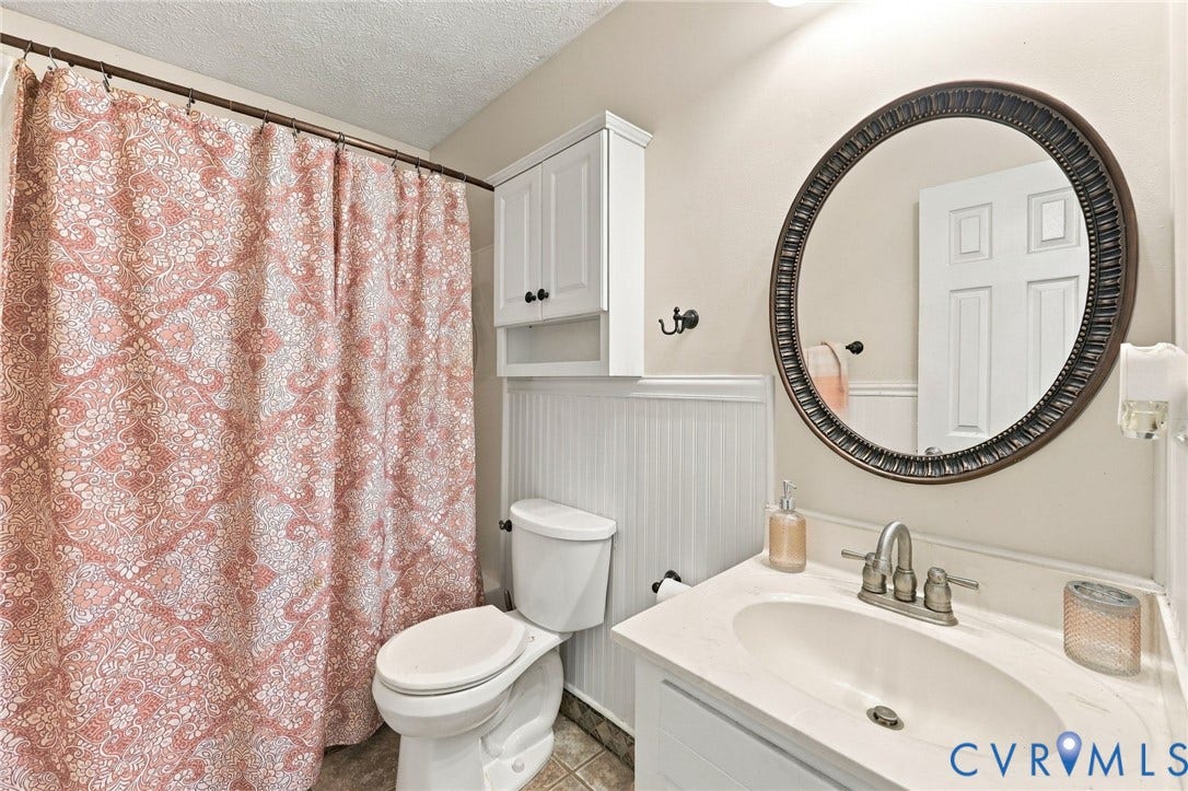 Bathroom featuring a white vanity with a neutral solid-surface countertop, an oval wall mirror with a dark metallic frame, wainscot paneling, and an upper built-in storage cabinet