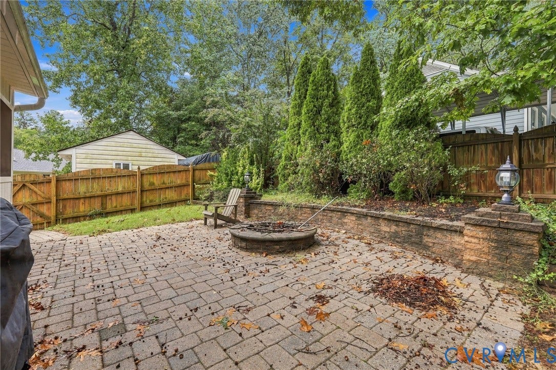 Paver patio featuring a circular stone fire pit, tiered retaining wall, wood privacy fencing, and mature evergreen landscaping