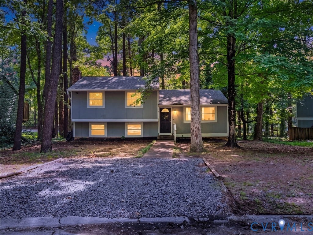 2703 Sutters Mill Court, Sutters Mill, Chesterfield, VA 23112 Split-level exterior featuring grey siding, a front-facing brick chimney, concrete walkway, gravel parking area, and mature perimeter trees