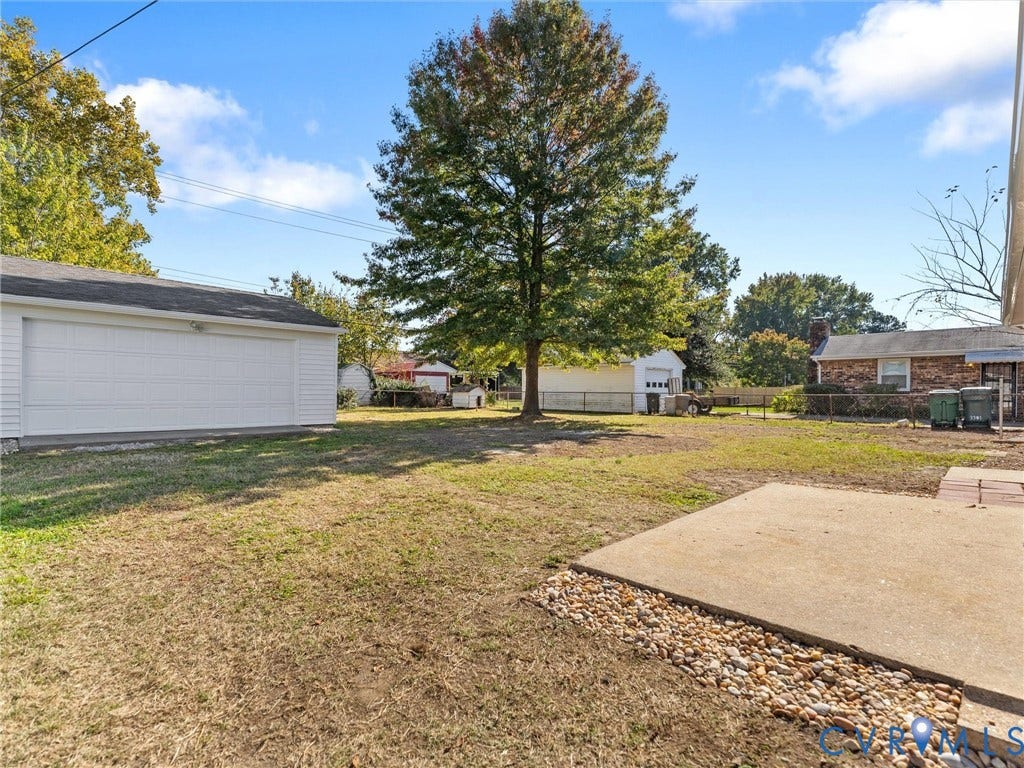 3303 Jowin Lane, Henrico, VA 23223 Rear exterior featuring a concrete patio, stone perimeter, detached garage, and mature trees within a fenced yard