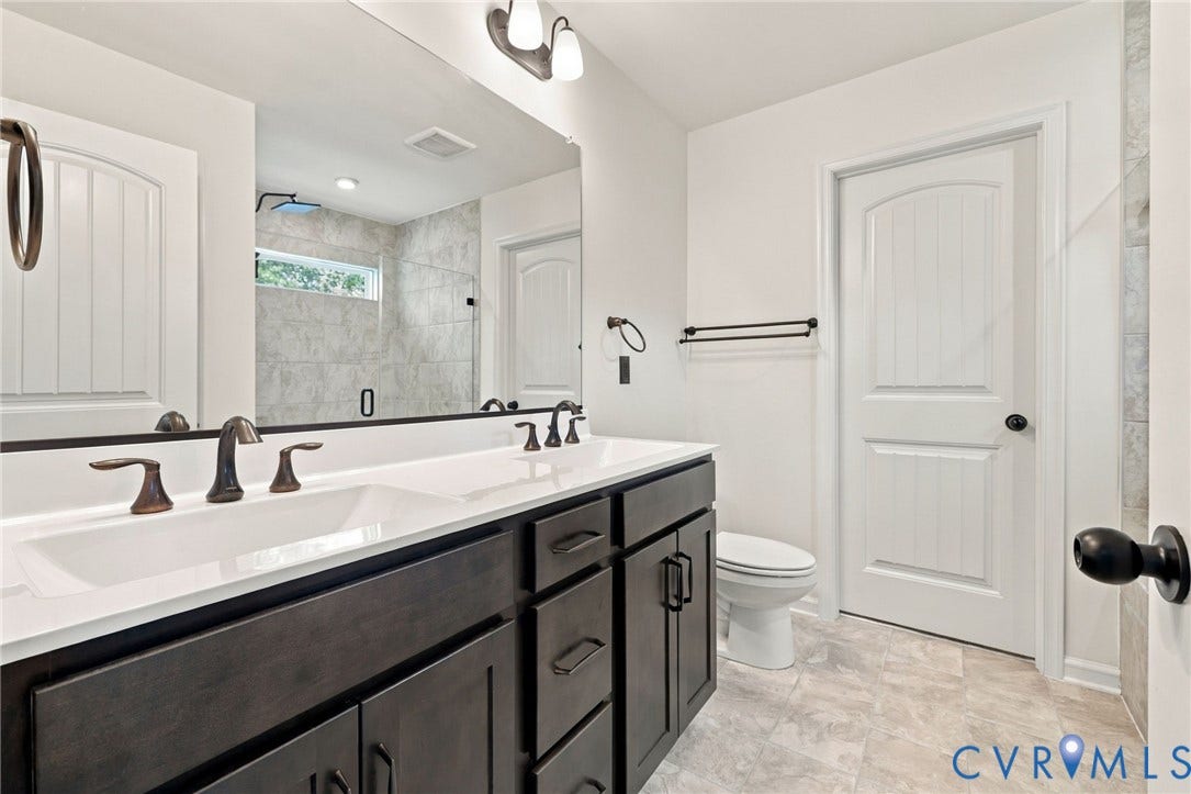 7712 Hawthorne Avenue, Henrico, VA 23227 Dual-sink vanity with dark cabinetry and composite countertop, large wall-mounted mirror, matte black fixtures, and tiled walk-in shower