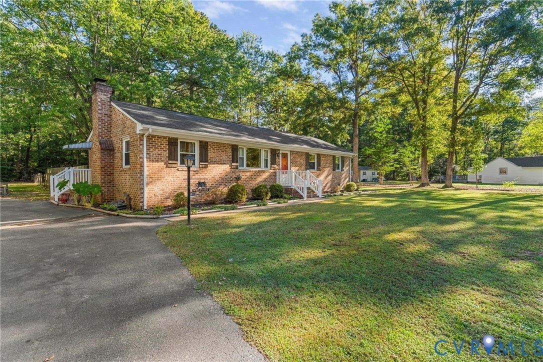 8444 New London Road, Mechanicsville, VA 23116 Brick exterior featuring a masonry chimney, dark-toned shutters, a paved driveway, and white railing accents, all situated on a lot with mature trees