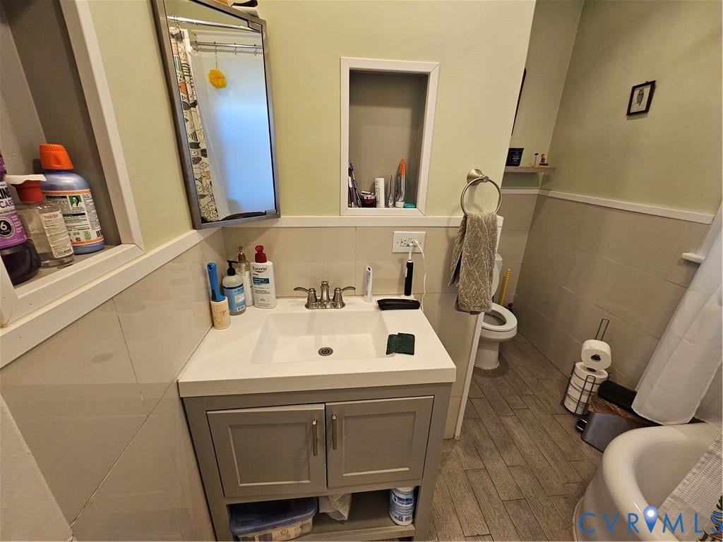 1206 Westminster Avenue, Richmond, VA 23227 Bathroom featuring a vanity with a white integrated sink, polished metal faucet, inset wall shelving, partial wall tiling, and wood-finish flooring