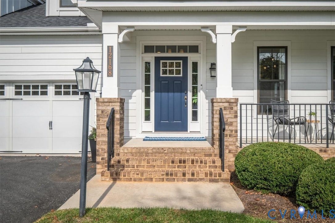 12168 Readers Pointe Drive, Readers Branch, Goochland, VA 23103 Exterior entry featuring horizontal siding, a covered porch with brick foundation piers, a solid-color paneled door with sidelights and transom window, and a carriage-style garage door