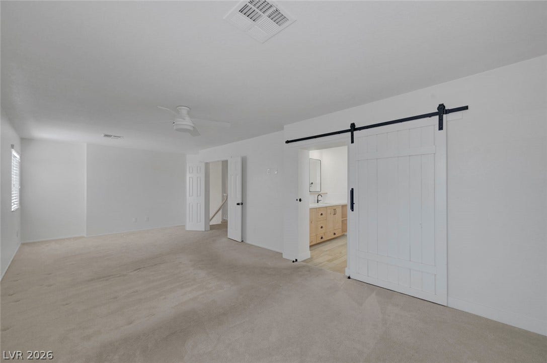 31 Antero Drive, Henderson, NV 89074 A spacious primary bedroom features soft carpeting, a ceiling fan, and a rustic barn door leading to an en-suite bathroom.