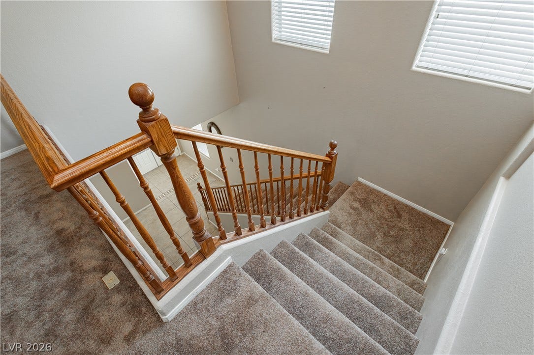 1176 Via Piazza, Henderson, NV 89052 A grand wooden staircase with carpeted steps descends into a tiled entryway, illuminated by high-set windows.