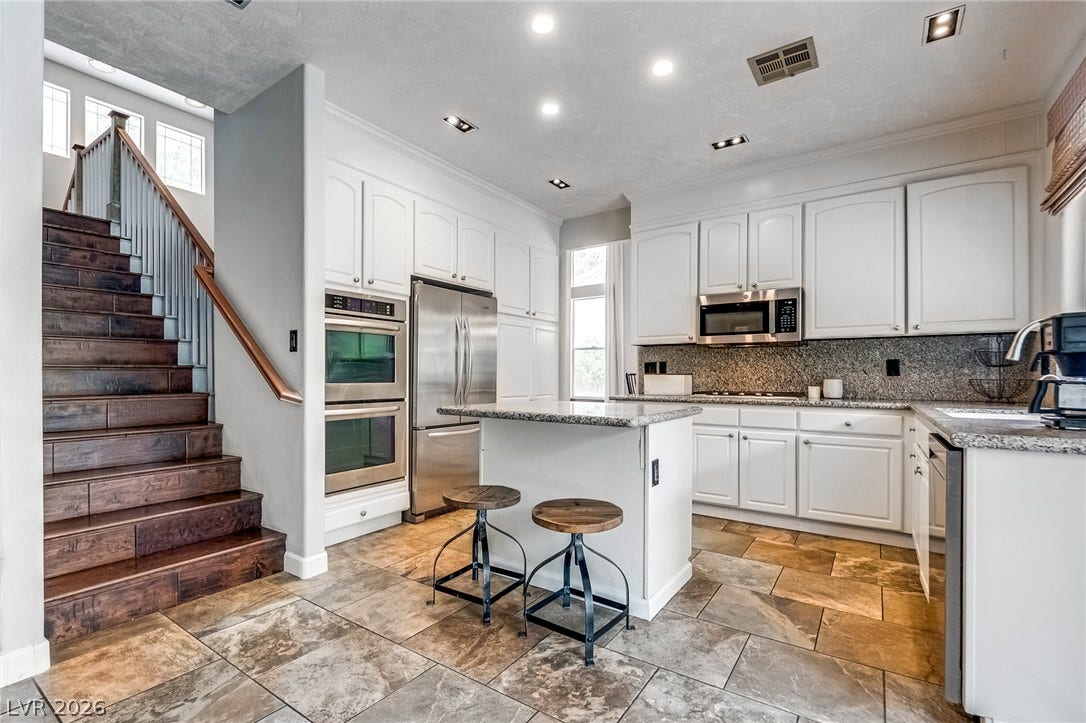 2393 Stansbury Court, Henderson, NV 89052 Bright white cabinetry and stainless steel appliances adorn this kitchen with a granite island, complemented by stone flooring and a rich wooden staircase.