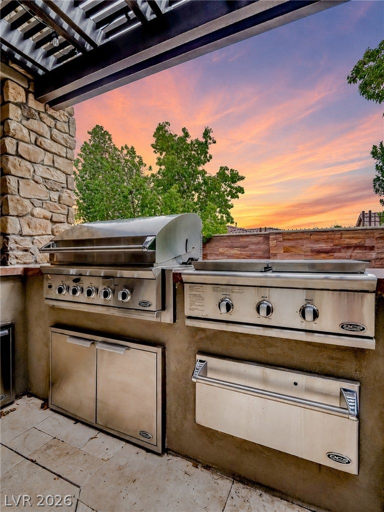 12013 Tramonto Avenue, Las Vegas, NV 89138 An outdoor kitchen features premium stainless steel appliances beneath a structural pergola at sunset.
