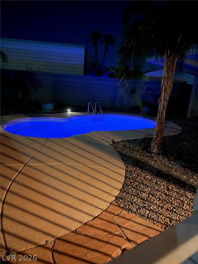 An illuminated in-ground pool beckons for evening relaxation with surrounding hardscaping and mature landscaping.
