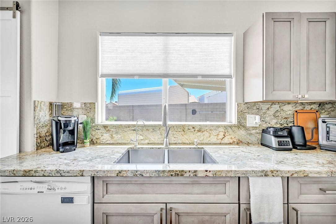 A well-lit kitchen presents a double basin stainless steel sink and light granite countertops.