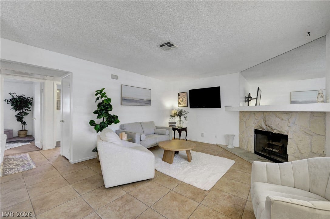 A well-lit living space features tiled flooring leading to a cozy seating area and a robust stone fireplace.