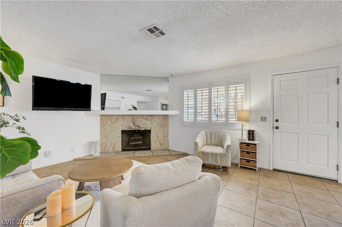 A corner stone fireplace anchors this bright living space, complemented by a large window with white shutters.