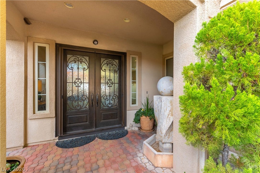 2676 Botticelli Drive, Henderson, NV 89052 Ornate wrought iron and glass double doors open to a welcoming entryway, framed by a serene water feature and vibrant landscaping.