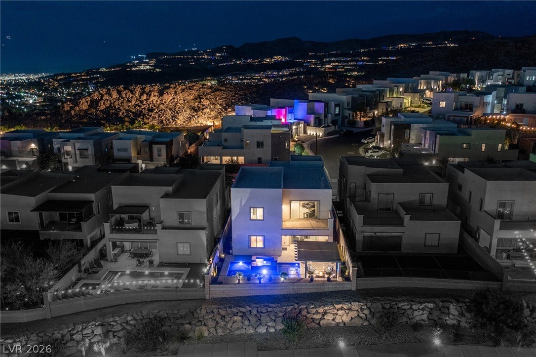 743 Glowing Horizon Street, Henderson, NV 89052 An aerial perspective reveals a modern home with a vibrant blue-lit pool and patio, set against a twinkling city hillside at dusk.