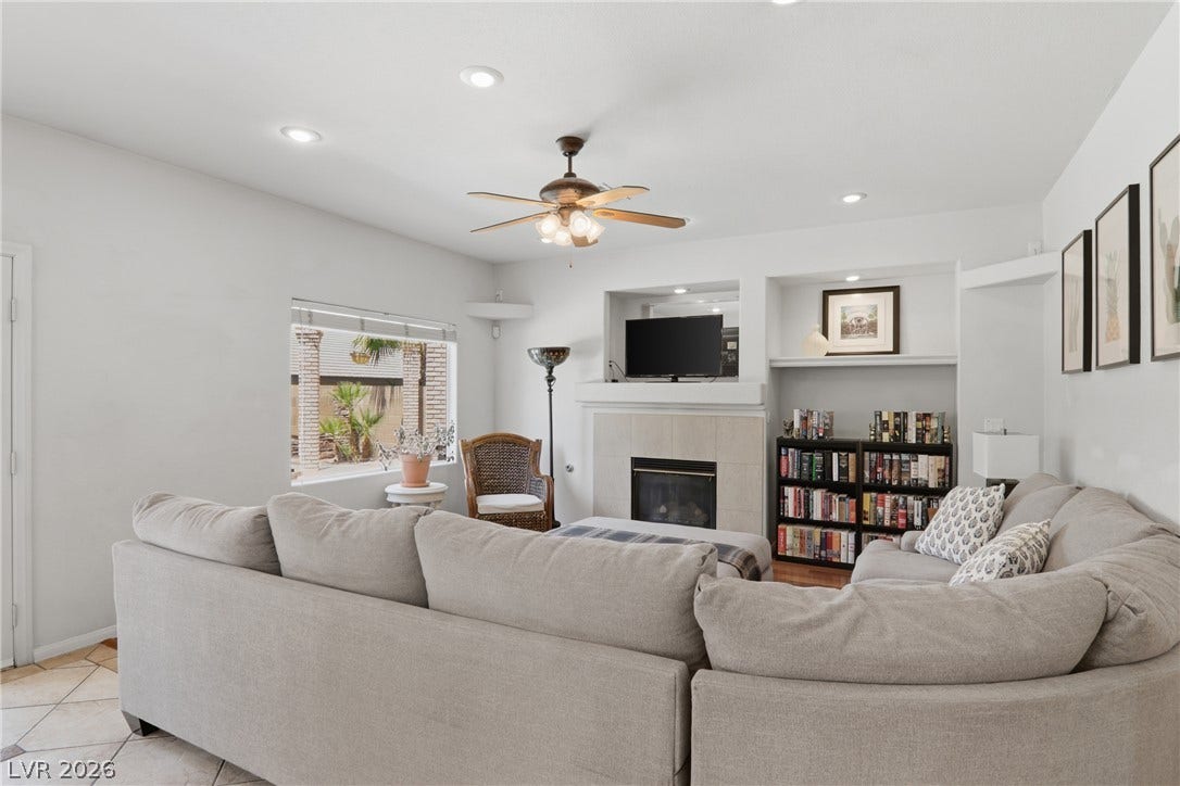 2218 Alanhurst Drive, Henderson, NV 89052 A comfortable living area features a corner fireplace, built-in shelving, and a window framing lush outdoor greenery.