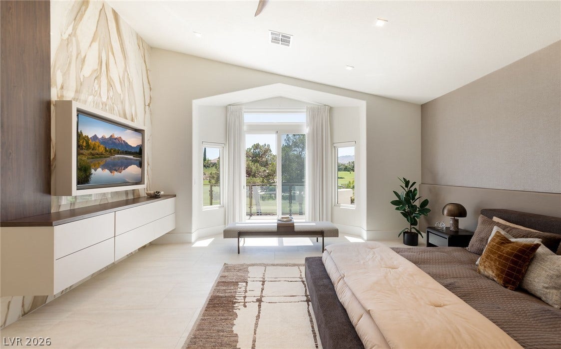A tranquil primary bedroom features an elegant stone accent wall, high ceilings, and a bay window framing serene outdoor vistas.