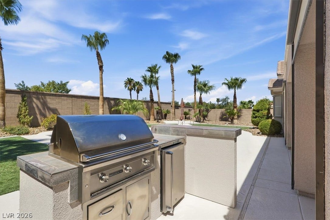 An outdoor kitchen features a built-in stainless steel barbecue grill against a palm tree backdrop.