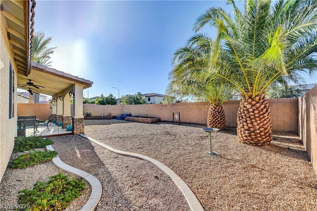 9928 Tradition Creek Avenue, Las Vegas, NV 89149 A spacious, rock-covered backyard features a covered patio with ceiling fans and two prominent palm trees.