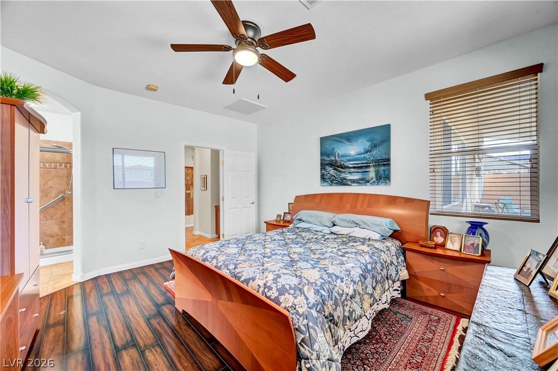 9928 Tradition Creek Avenue, Las Vegas, NV 89149 Rich hardwood flooring complements a spacious bedroom featuring a ceiling fan and natural light from a window with blinds.