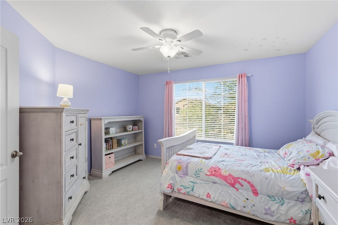 A well-lit bedroom with soft purple walls, plush carpeting, and an overhead ceiling fan.