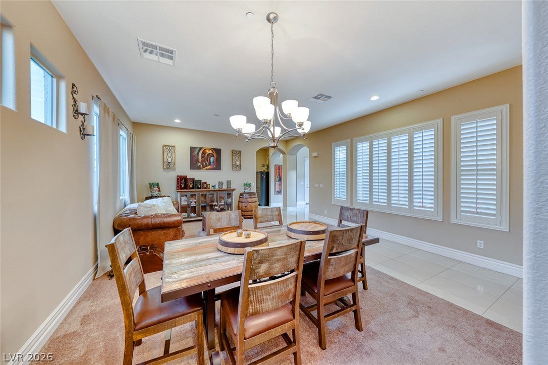70 Portezza Drive, Henderson, NV 89011 A rustic dining table anchors this open-concept space, illuminated by an elegant chandelier and natural light from shuttered windows.