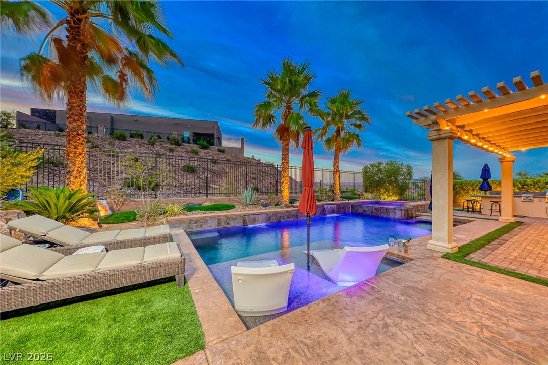 70 Portezza Drive, Henderson, NV 89011 An inviting backyard oasis features a dazzling pool with submerged lounge chairs, complemented by a covered patio perfect for evening entertaining.