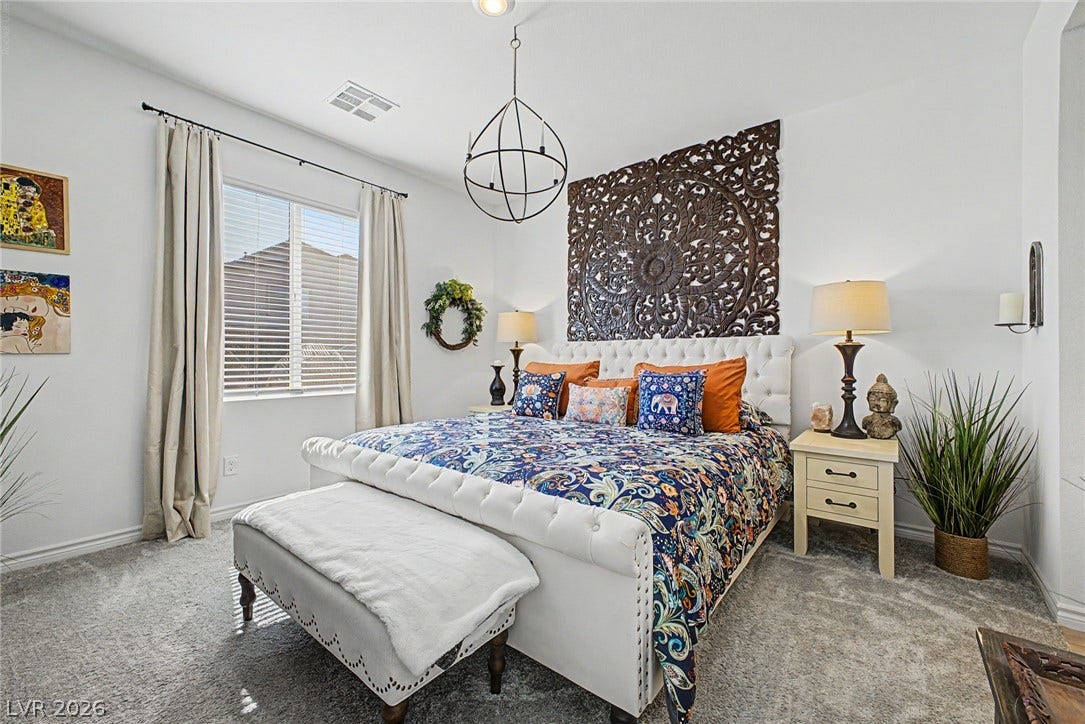 290 Fossil Falls Street, Henderson, NV 89015 A tranquil bedroom features plush carpeting, a wide window with blinds, and a distinctive chandelier overhead.