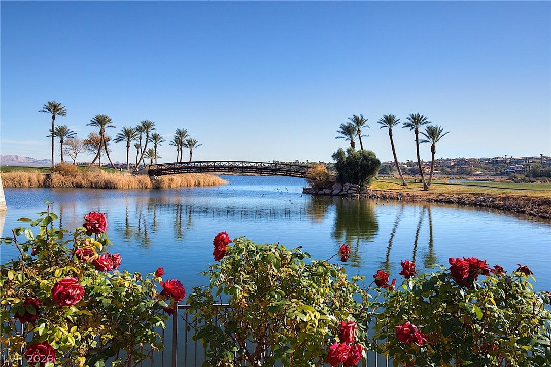 30 Benevolo Drive, Henderson, NV 89011 Flowering bushes frame a serene lake, reflecting palm trees and a charming pedestrian bridge.