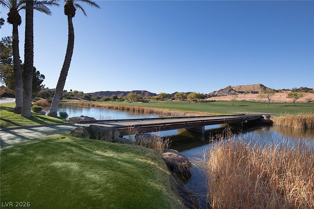 30 Benevolo Drive, Henderson, NV 89011 A tranquil golf course vista unfolds, featuring a serene pond, elegant bridge, and lush fairways backed by distant mountains.