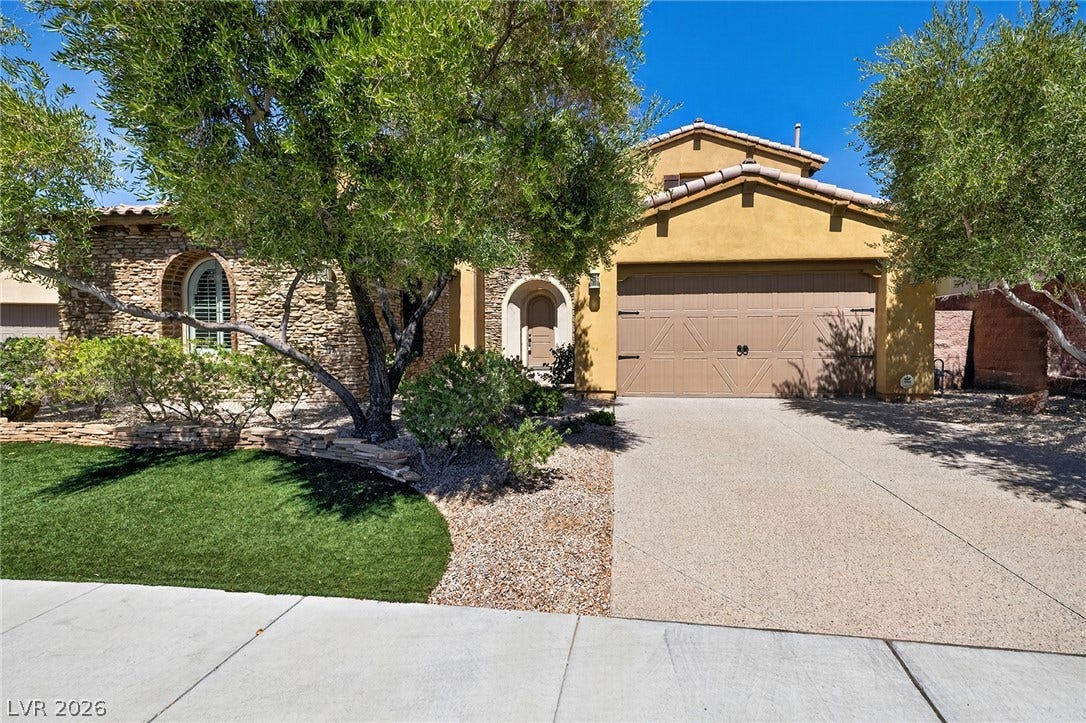 30 Benevolo Drive, Henderson, NV 89011 A welcoming stone facade home features an arched entry, mature landscaping, and a driveway leading to a two-car garage.
