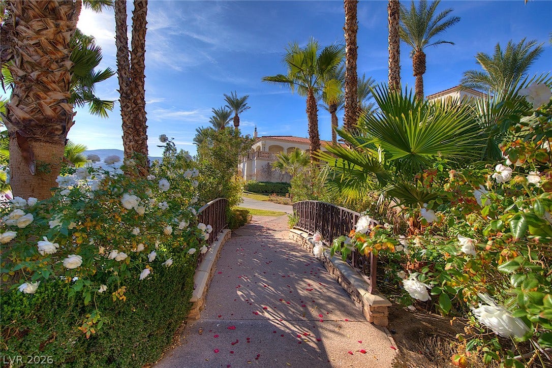 30 Benevolo Drive, Henderson, NV 89011 A charming footbridge winds through lush landscaping, leading to a majestic residence under a brilliant sky.