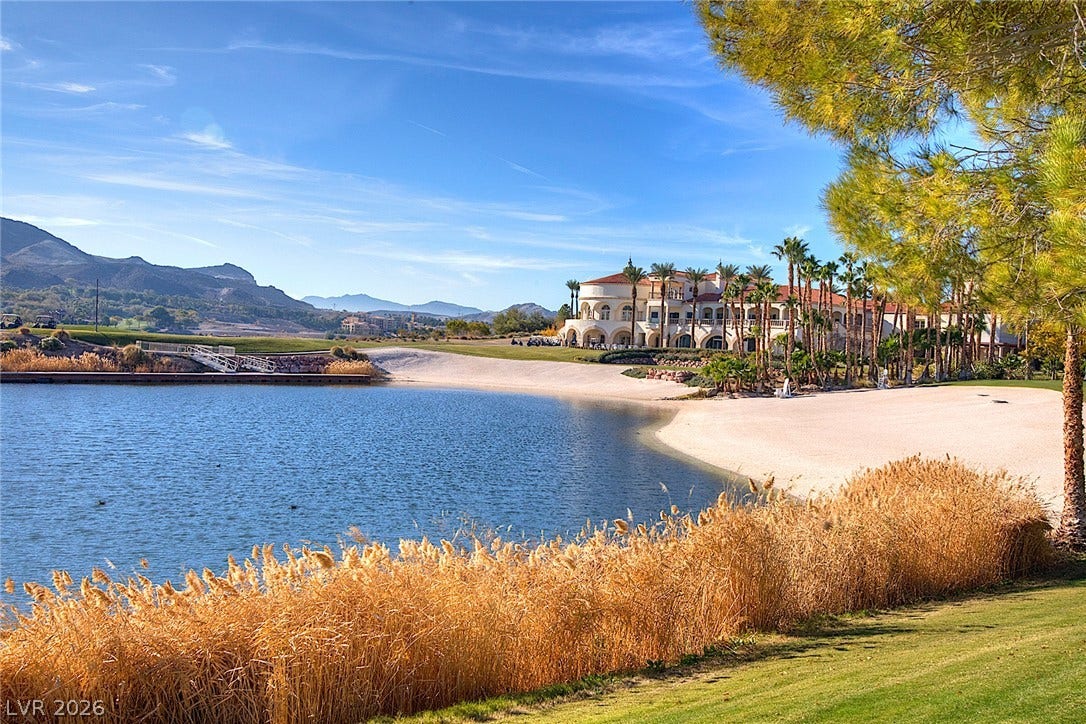 30 Benevolo Drive, Henderson, NV 89011 An exquisite golf course landscape unfolds, featuring a sparkling lake, sandy beach, and a grand Mediterranean-style clubhouse nestled against a mountain backdrop.
