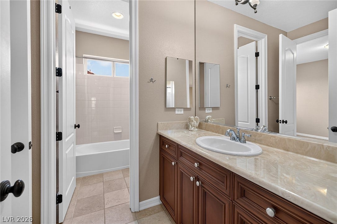 30 Benevolo Drive, Henderson, NV 89011 A well-appointed bathroom features a long vanity with an integrated sink and warm wooden cabinetry, complementing the tiled flooring and bathtub.