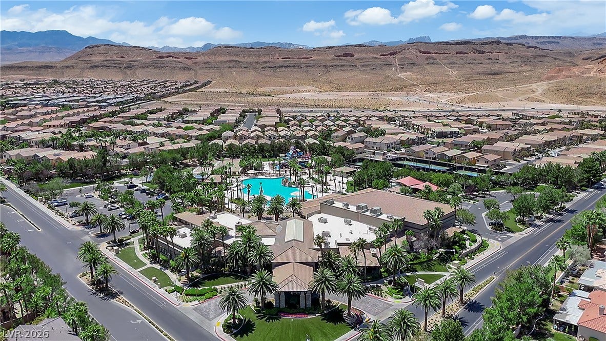 771 White Dogwood Court, Las Vegas, NV 89148 An aerial panorama reveals a vibrant community hub with a sparkling pool and lush landscaping, nestled among residential homes against a dramatic desert mountain backdrop.