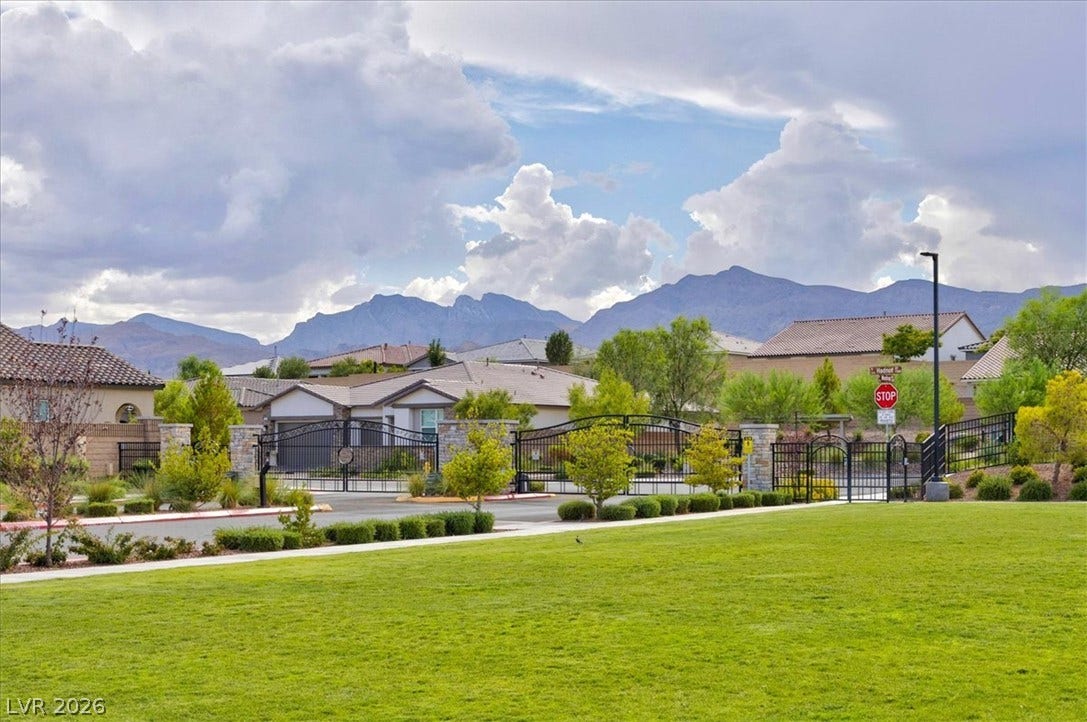 A manicured green lawn leads to a gated community entrance with majestic mountains framing the background.