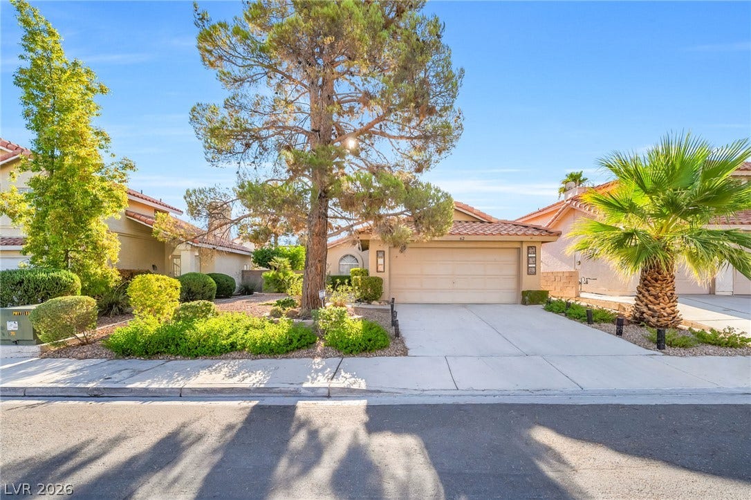 62 Tidwell Lane, Henderson, NV 89074 Mature trees frame the concrete driveway leading to a classic stucco home with tiled roofing.