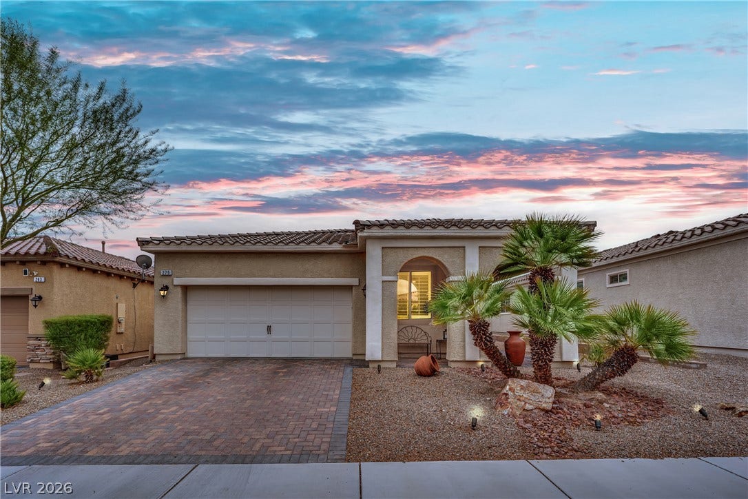 A well-maintained stucco home with a paved driveway and desert landscaping, set against a colorful sunset sky.