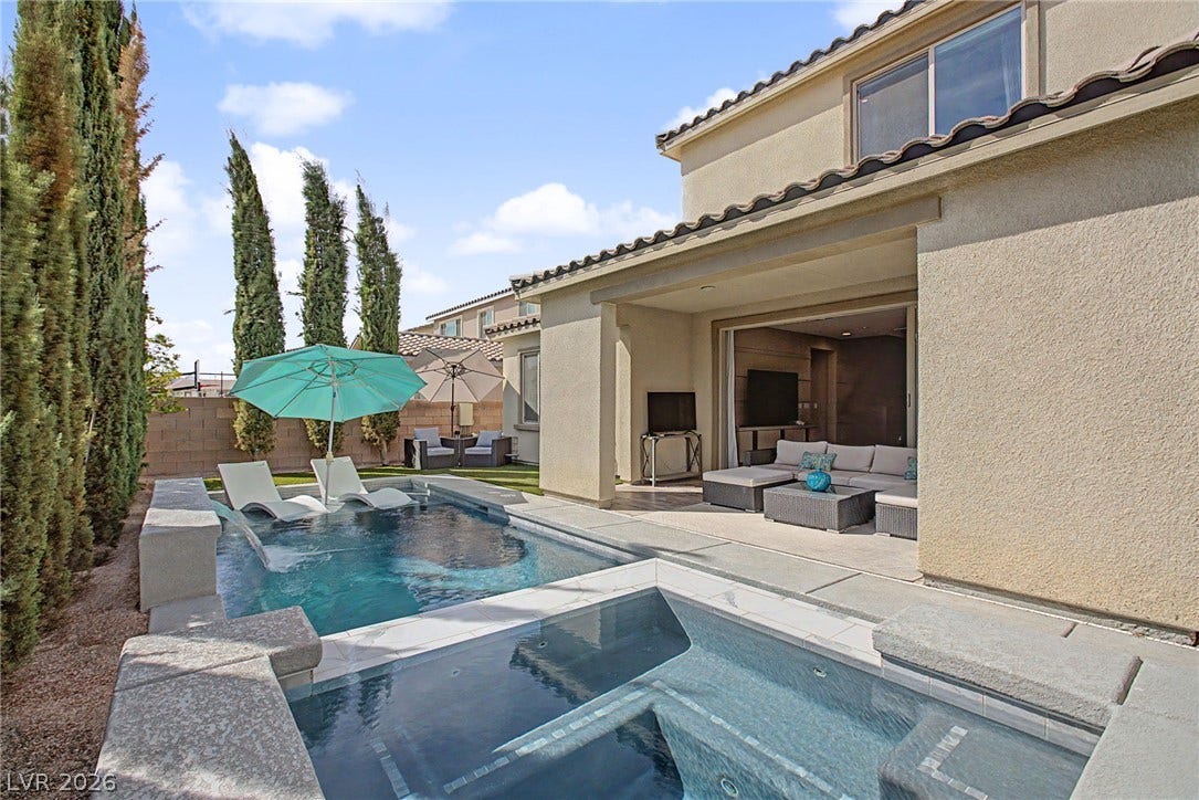 A modern pool and spa sit adjacent to a covered stone patio and stucco exterior.