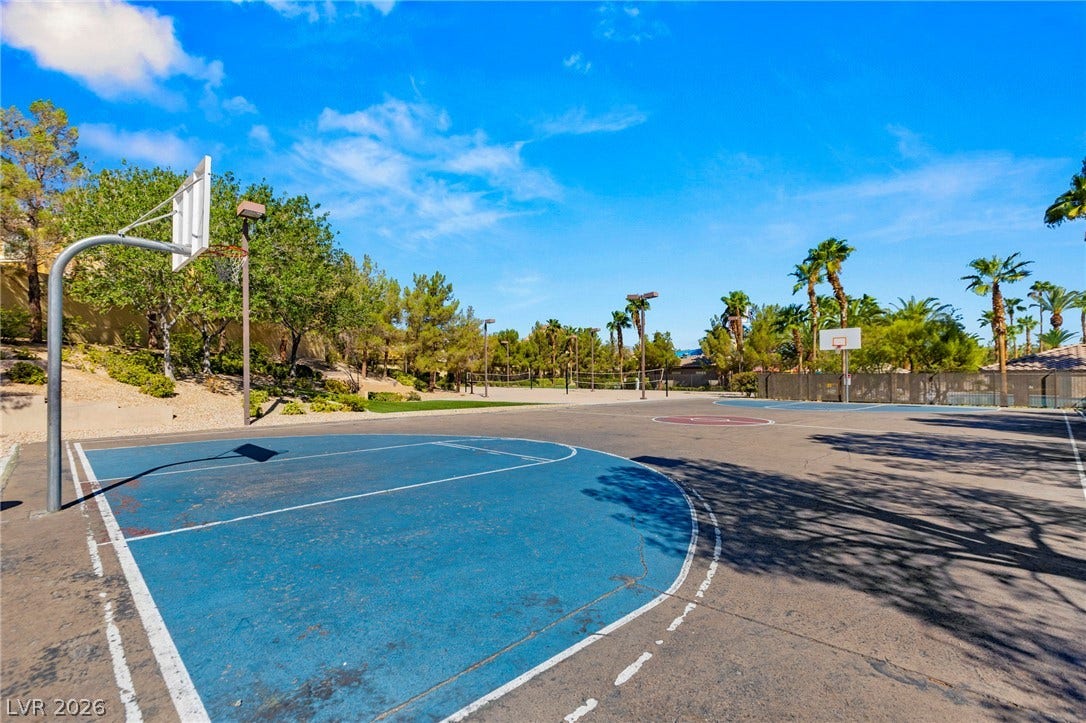 An outdoor basketball court stands surrounded by lush palm trees under a bright blue sky.