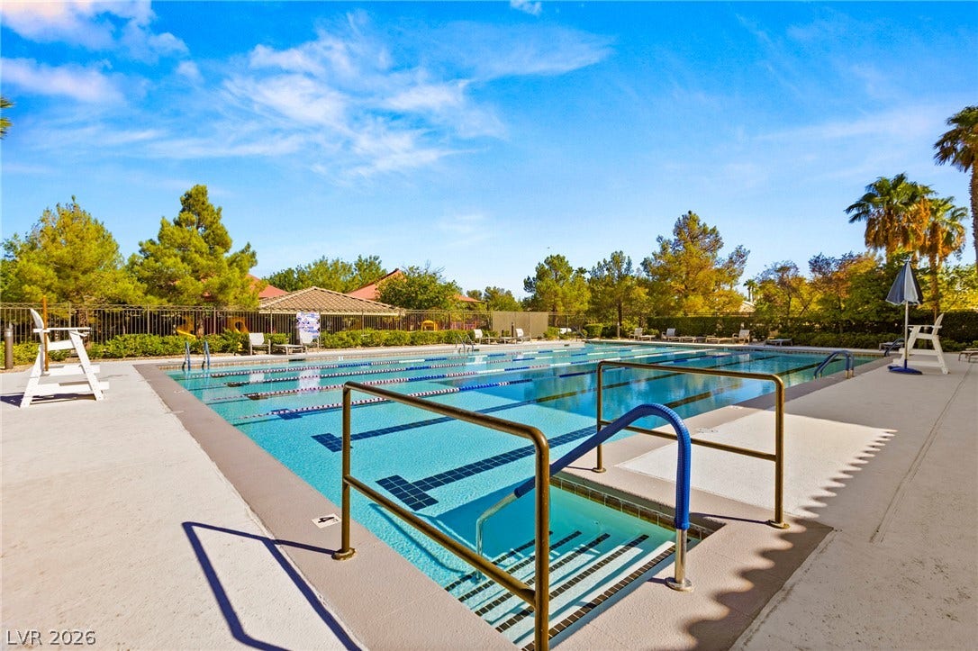 A spacious outdoor lap pool is bordered by concrete decking and lush green trees.