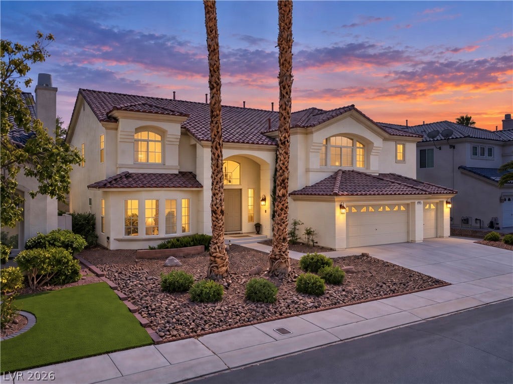 Evening view of the two-story estate showcasing its grand entry, three-car garage, towering palm trees, and mature landscaping.